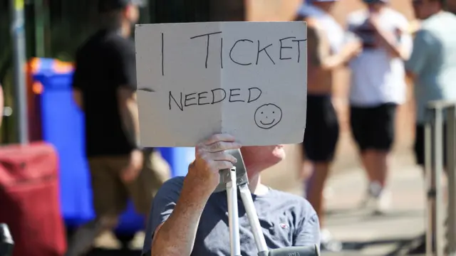A man in a blue T-shirt and holding crutches carries a sign covering his face which reads: 1 Ticket Needed