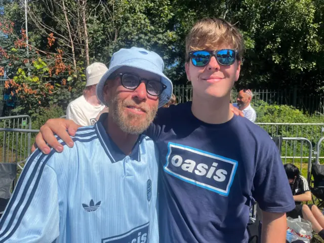 The two fans are both wearing blue Oasis T-shirts and are smiling at the camera.