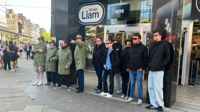 A number of men are standing in front of Primary wearing black and green parka jackets