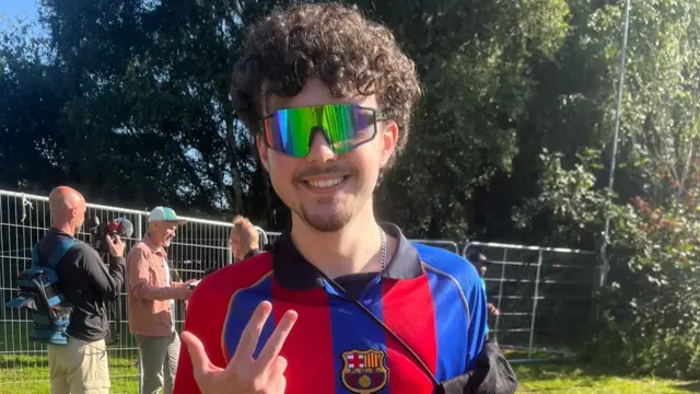 Theo is wearing sunglasses and a Barcelona T-shirt. He is standing outside Heaton Park and he is smiling at the camera