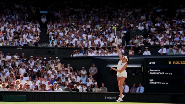 Iga Swiatek during her Ladies' Singles Final reaches racket into air to hit ball at final match, with a crowd of spectators watching on.
