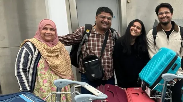 Four people, a woman, man, woman, man, smiling and holding each other in a line in front of suitcases piled up on trolleys