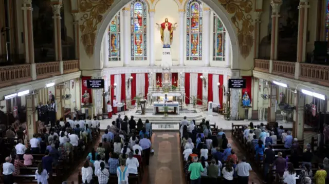 Taken from above, the picture shows the view of inside a church behind the people attending. The pews are full and they look towards the altar, which is elaborate and set up with candles and a statue of Jesus