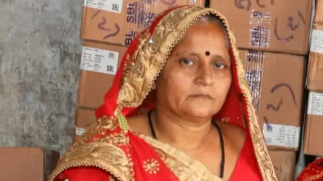 A woman wearing a red saree looks into the camera. There are boxes in the background.