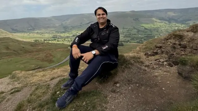 A man sat on the ground on top of a high hill with hills in the distance. He smiles at the camera