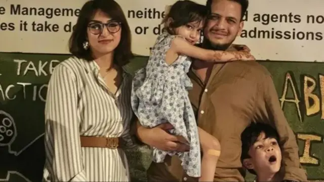 A mum and dad smiling as the dad holds a young girl who clings to his neck, and his son stands next to them with his mouth open