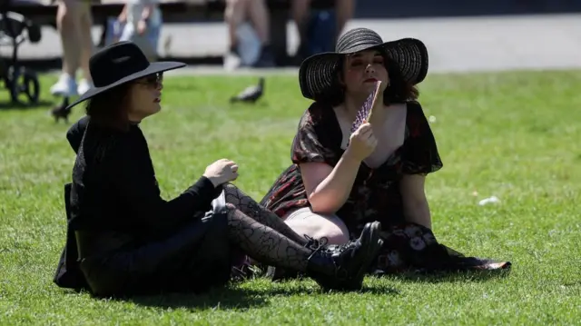 Two women sit on the grass with hats and fans
