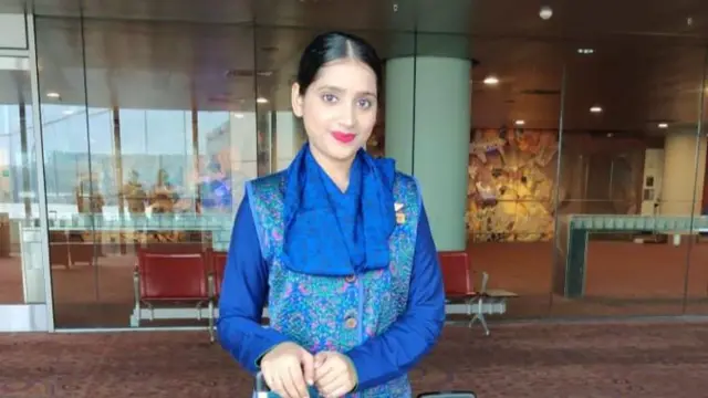 A woman smiling at the camera as she stands in an airport with a bright blue outfit on and pink lipstick