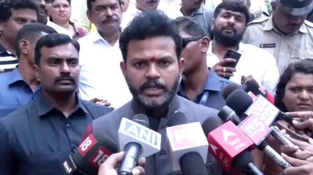 A man speaking into several press microphones that are being held to his face as he is crowded by people all around him. He looks serious as he talks