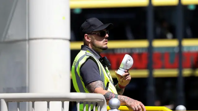 A man holds a personal fan and wears a high-vis vest and a cap.
