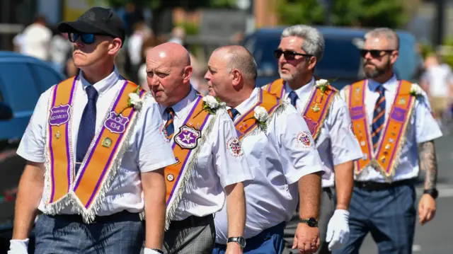 Men stand in a line, wearing white shirts, ties and orange sashes.