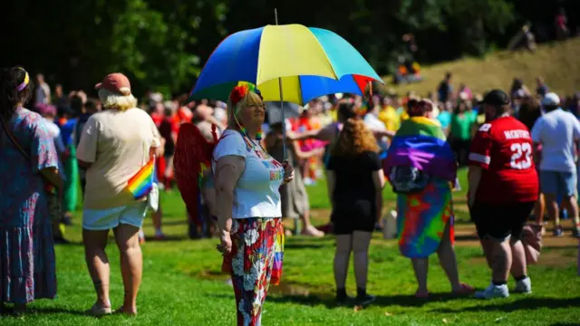 A woman wears a colourful outfit and holds a colourful umbrella with a crowd of people behind her.