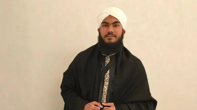 A man wearing a white turban and a black cape with a black shirt and golden embroidery on it.