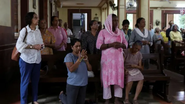 A group of people in a church, some sit and some stand. They look sad and mournful as they look ahead of them