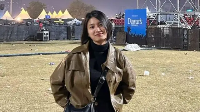 A woman wearing a brown jacket stands in front of a concert ground