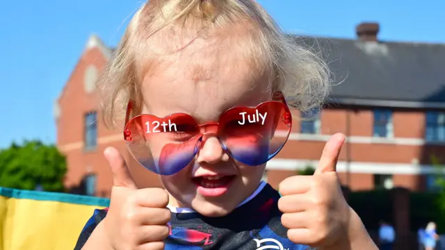A girl looks ahead, with both thumbs up, wearing big glasses that have 12th July written on them.