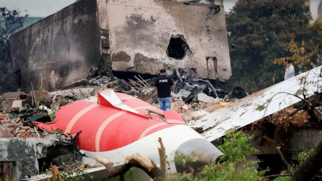A fire fighter stands on top of the wreckage of the crashed plane, which is in pieces and among rubble of a building. There's a large concrete block in front of the wreckage