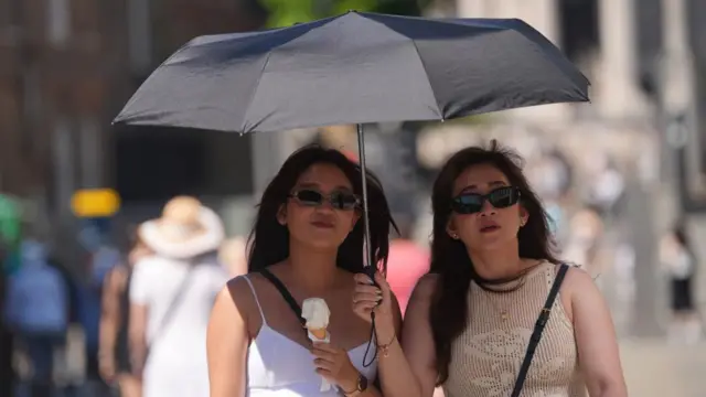Two women shelter from the sun under a black umbrella - one holds an ice cream