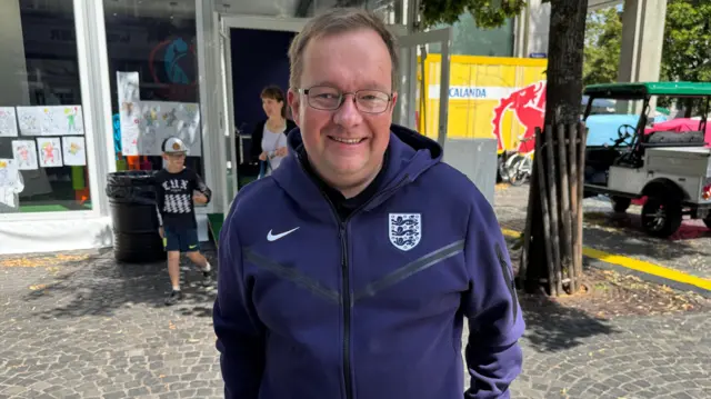 England fan Phillip Spakouskas. He is wearing a navy blue zipped jumper with the Coat of Arms of England on it, and has short brown hair and glasses. He is standing on a cobbled street with a tree and family behind him.