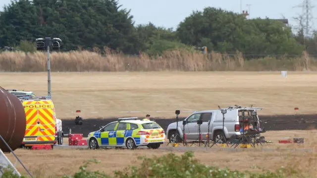 Three vehicles at Southend Airport on a grassed area where a plane crashed. A wide area of blackened grass is surrounded by a brown, grassy area.