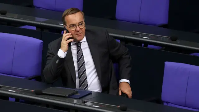 Boris Pistorious sits amongst a group of empty purple chairs, speaking on a mobile phone.