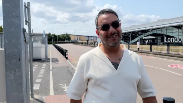 A man in a white top, wearing shades, standing next to a road in front of the London Southend Airport terminal building