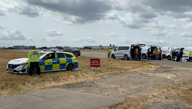 A police car and six other vehicles on a runway and grass verges at an airport. A red sign says "CAUTION; ENGINE TEST AREA AHEAD"