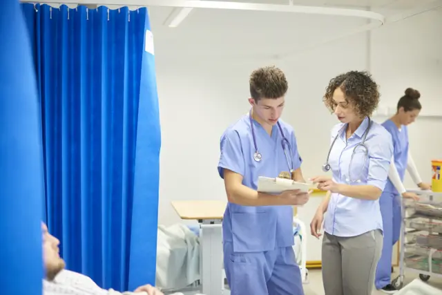 Two doctor's in a hospital ward. One is holding at clipboard which both doctors are looking at.