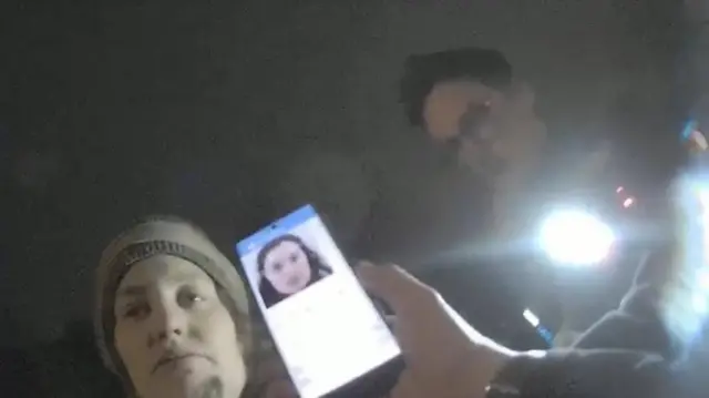Police bodycam image from the perspective of an officer holding a mobile phone displaying a picture of Constance Marten alongside Constance Marten herself. Behind her another officer stands with a bright torch
