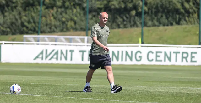Liam Manning, wearing Norwich City training kit, walks across the pitch with a football in the foreground