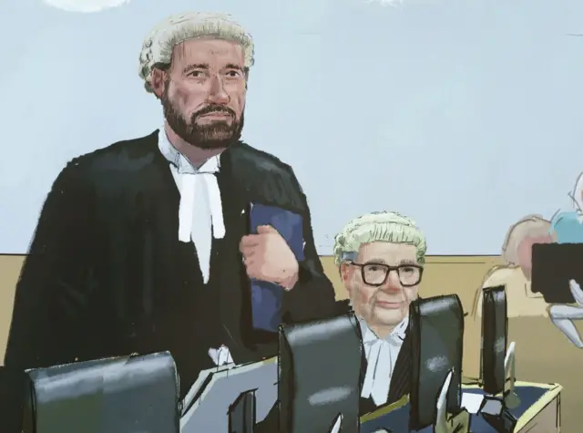 Andrew Gurney, who has a neat dark beard and is wearing a cream wig and black robes stands holding a folder to his chest next to Christopher Knox, who is seated and had glasses and a cream wig. A row of computer screens are in front of them.
