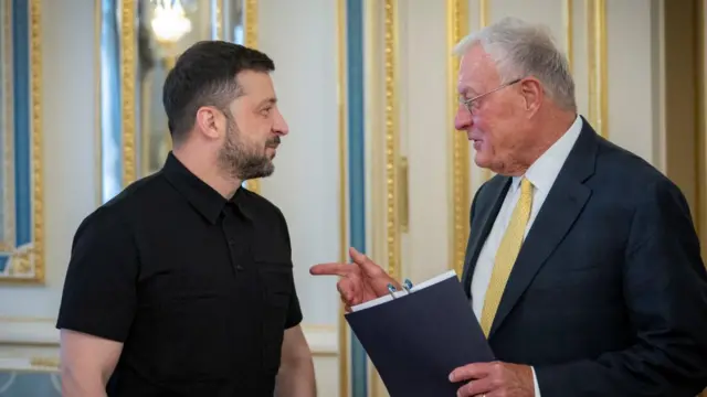 Zelensky speaking with Keith Kellogg. Kellogg is holding a folder and pointing toward Zelensky