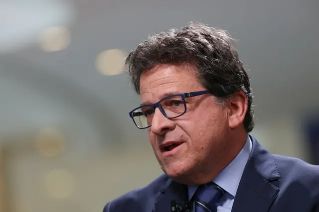 Mark Attanasio, pictured wearing a blue suit, light blue shirt and blue diagonal-striped tie. He has black hair and is wearing black-framed glasses.