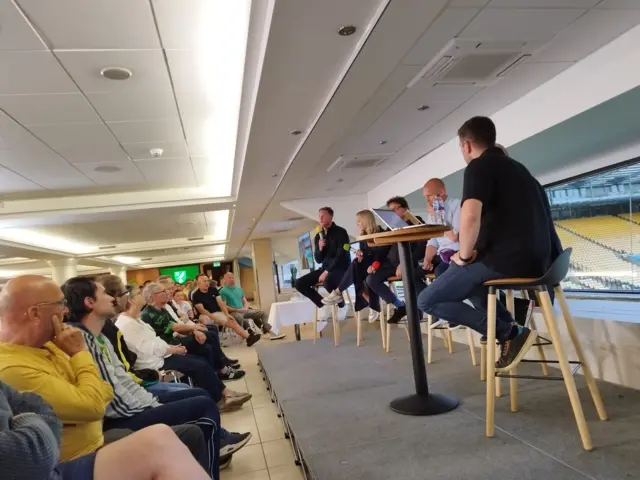 A crowd listens to people speaking on a stage with microphones.