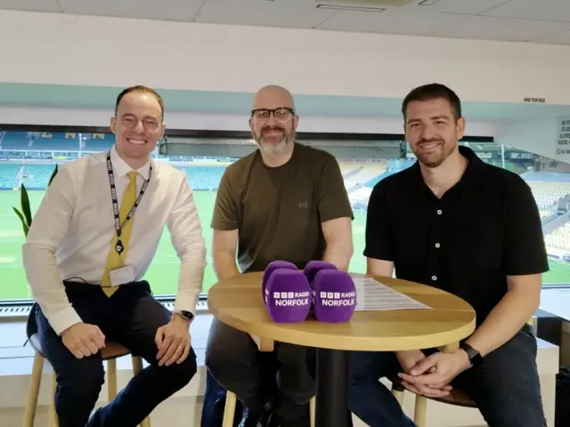Three male radio presenters sit on high stools around a table ahead of a live event at a football stadium, with the pitch seen behind them through windows