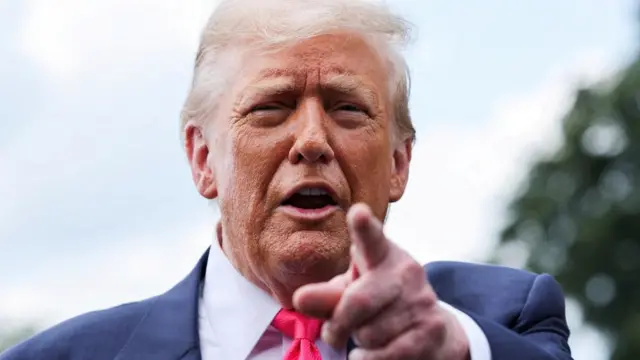 Trump pointing at the camera with his hand while speaking. He appears to be outside but not much background is visible, just a blurred tree to one side.