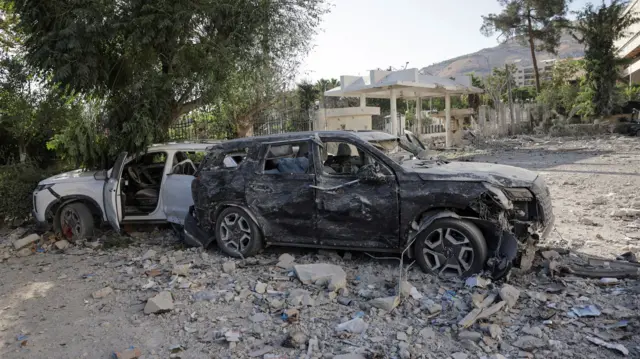 A black damaged car facing right and and behind it, a white damaged car facing left with the passenger door open. Both cars are sitting on debris