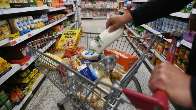 A man places a carton of milk into a supermarket trolley which is full of household staples like sliced bread, cereal, butter and pasta