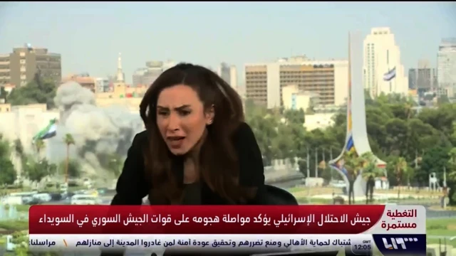 A still image from a Syrian TV broadcast showing a news anchor in a studio reacting to strike on a building behind her. She is sat in a chair and appears to be bracing as smoke rises from a building behind her.