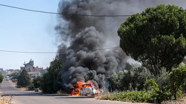 Smoke billows from a burning car on the side of an empty road