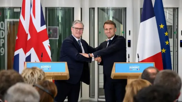 Starmer and Macron standing at two podiums with microphones on them that read UK France Summit on the front. Behind them are EU, British and French flags. They smile facing forwards while putting their arms on each other in an embrace. Rows of people sit in front of them with the back of their heads in focus.