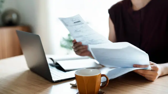 Woman sitting at dining table, handling personal finance papers with laptop and mug.
