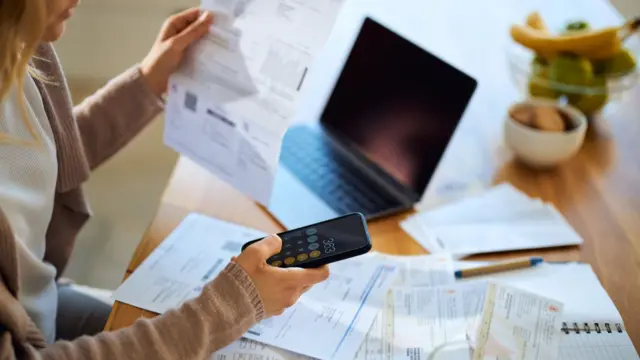 Unrecognizable woman using mobile phone while calculating the amount of her bills at home. Focus is on hand and cell phone.