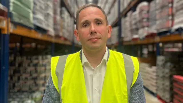 Neil Gamble wears a hi vis jacket in front of pallets of food in a warehouse