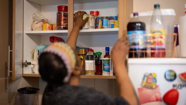 A woman, wearing a headband and t-shirt, reaches into a kitchen cupboard which contains tins of fish, chickpeas and other items