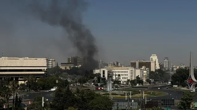 A city view taken from what seems like on top of high ground. There's a large roundabout with a fountain in the middle, and black smoke rises behind it from a building