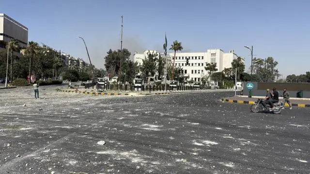 A zoomed out picture of the building where you can see white dust across the road and bits of rubble as people drive by and stand there