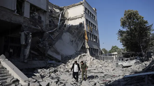 Two men walking among rubble. behind them is a damaged building.