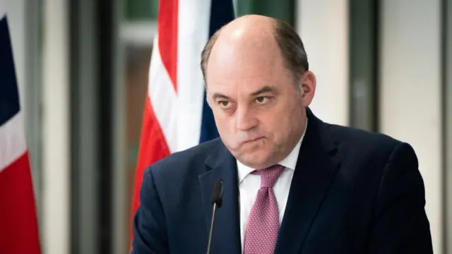 Headshot of Ben Wallace looking concerned with his mouth shut tight as he stands in front of a small microphone and two British flags behind him