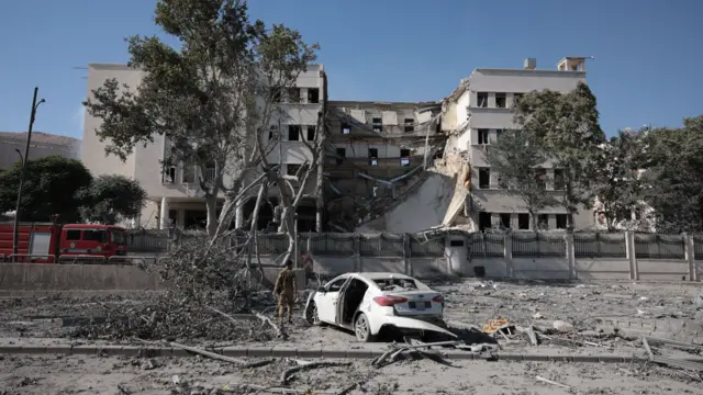 A building that has been damaged at the centre. In front of it is debris and a damaged white car. There are three trees in the foreground